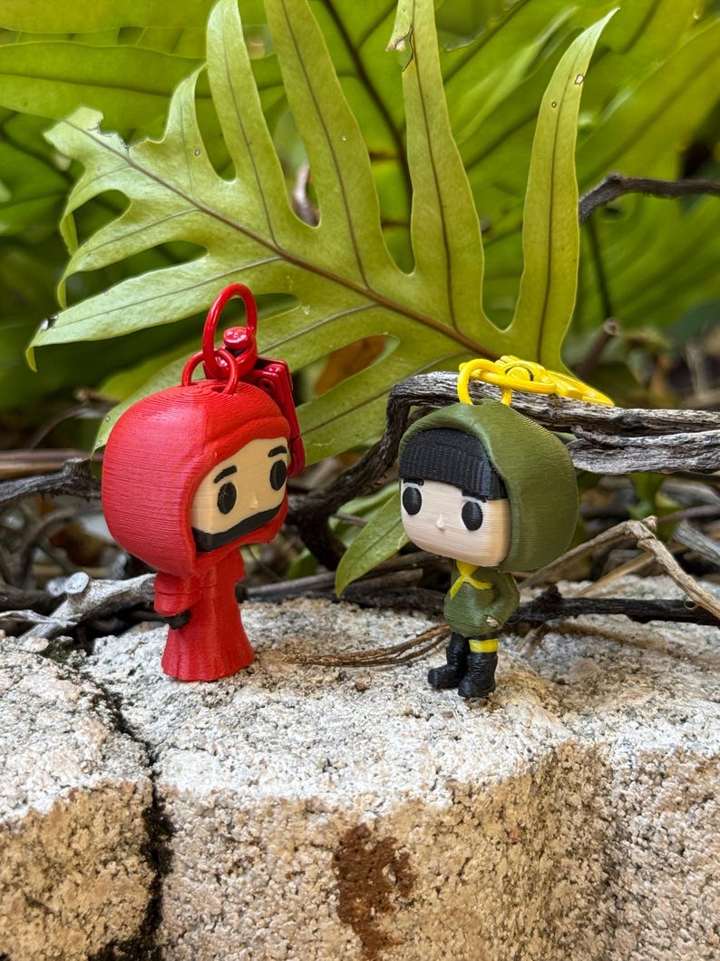 May include: Two miniature figurines, one red and one green, stand side-by-side on a stone surface. The red figure wears a hooded robe, while the green figure is dressed in a military-style outfit. Both have black boots and yellow accents. Keychains are attached.