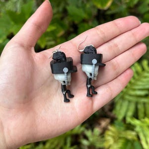 May include: A pair of black and white 3D printed earrings shaped like a person wearing a hat. The earrings are hanging from silver hooks.