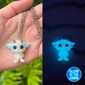 May include: A white glow-in-the-dark necklace with a small, white, cartoon character pendant with blue accents. The pendant is shaped like a creature with large eyes and ears. The necklace is on a silver chain. The text "GLOW IN THE DARK" is visible in the bottom right corner of the image.