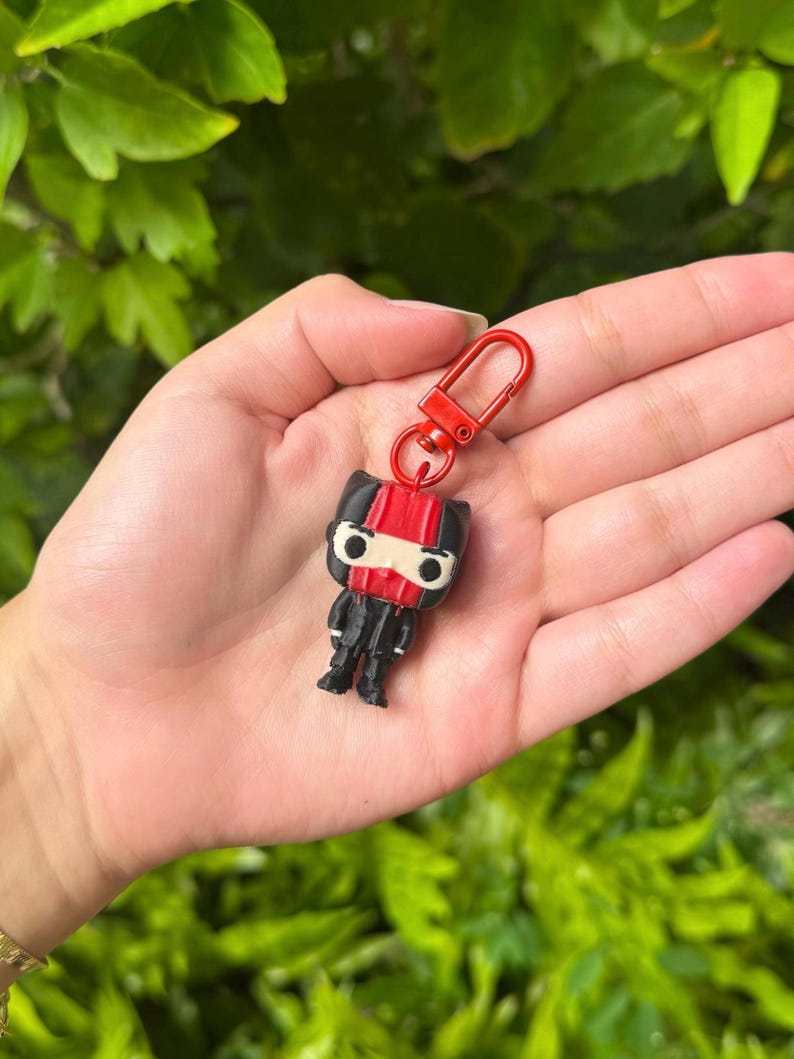 May include: A black and red character keychain with a red mask and a red and black striped top. The keychain is attached to a red metal ring.