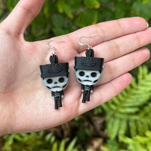 May include: A pair of silver skeleton earrings with black hats. The skeletons are wearing black and white uniforms.