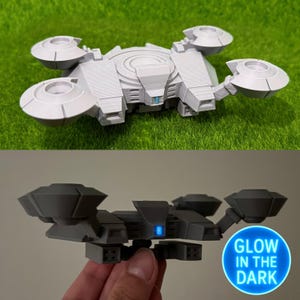 Może przedstawiać: Szary, drukowany w 3D model futurystycznego statku kosmicznego lub drona. Projekt zawiera wiele okrągłych i kątowych elementów, z centralnym korpusem i czterema obracającymi się elementami w kształcie dysku. Model ma szczegółową, teksturowaną powierzchnię i niebieskie światło.