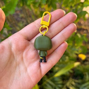 May include: A green figurine keychain with a yellow clasp. The figurine has black boots and yellow accents on the sleeves. The keychain is held in a hand against a blurred green background.