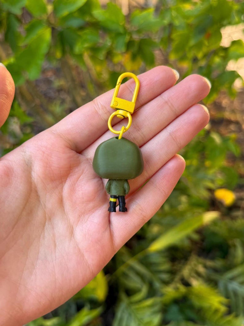 May include: A green figurine keychain with a yellow clasp. The figurine has black boots and yellow accents on the sleeves. The keychain is held in a hand against a blurred green background.