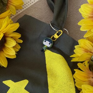May include: A dark green tote bag with a yellow graphic design and a small keychain of a cartoon character. The bag has a dark green strap and is surrounded by yellow sunflowers.