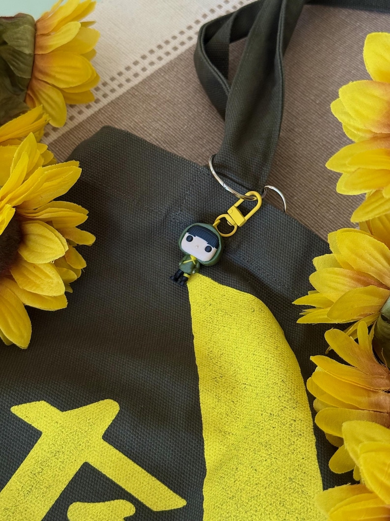 May include: A dark green tote bag with a yellow graphic design and a small keychain of a cartoon character. The bag has a dark green strap and is surrounded by yellow sunflowers.