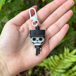 May include: Black and white skeleton keychain with a white hat and a white key ring. The keychain is in the shape of a person.