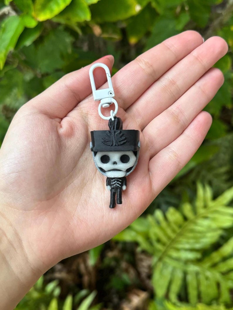 May include: Black and white skeleton keychain with a white hat and a white key ring. The keychain is in the shape of a person.