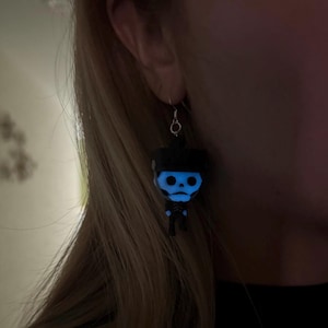 May include: A pair of glow-in-the-dark skeleton earrings. The earrings are black with a blue glow and feature a small skeleton figure.