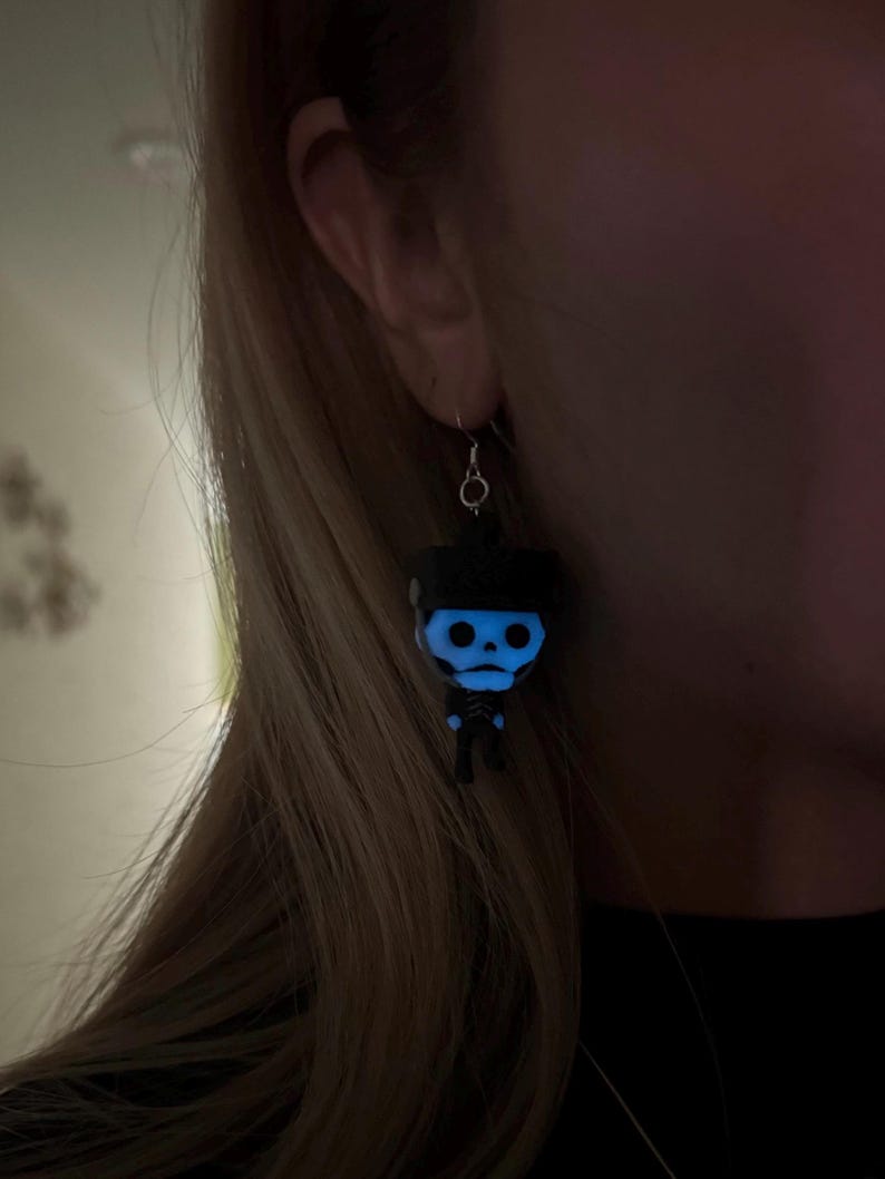 May include: A pair of glow-in-the-dark skeleton earrings. The earrings are black with a blue glow and feature a small skeleton figure.