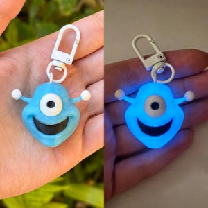 May include: A blue glow-in-the-dark keychain with a single white eye and a smiling mouth. The keychain is attached to a white metal clasp.