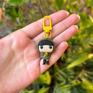 May include: A Funko Pop keychain featuring a figure in a green outfit with a black hood and black boots. The keychain has a yellow clasp and ring. The figure is held in a hand against a blurred green background.