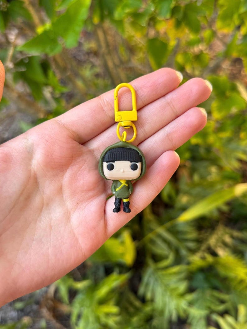 May include: A Funko Pop keychain featuring a figure in a green outfit with a black hood and black boots. The keychain has a yellow clasp and ring. The figure is held in a hand against a blurred green background.