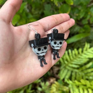 May include: A pair of black and white skeleton earrings with black hats. The earrings are hanging from silver hooks.