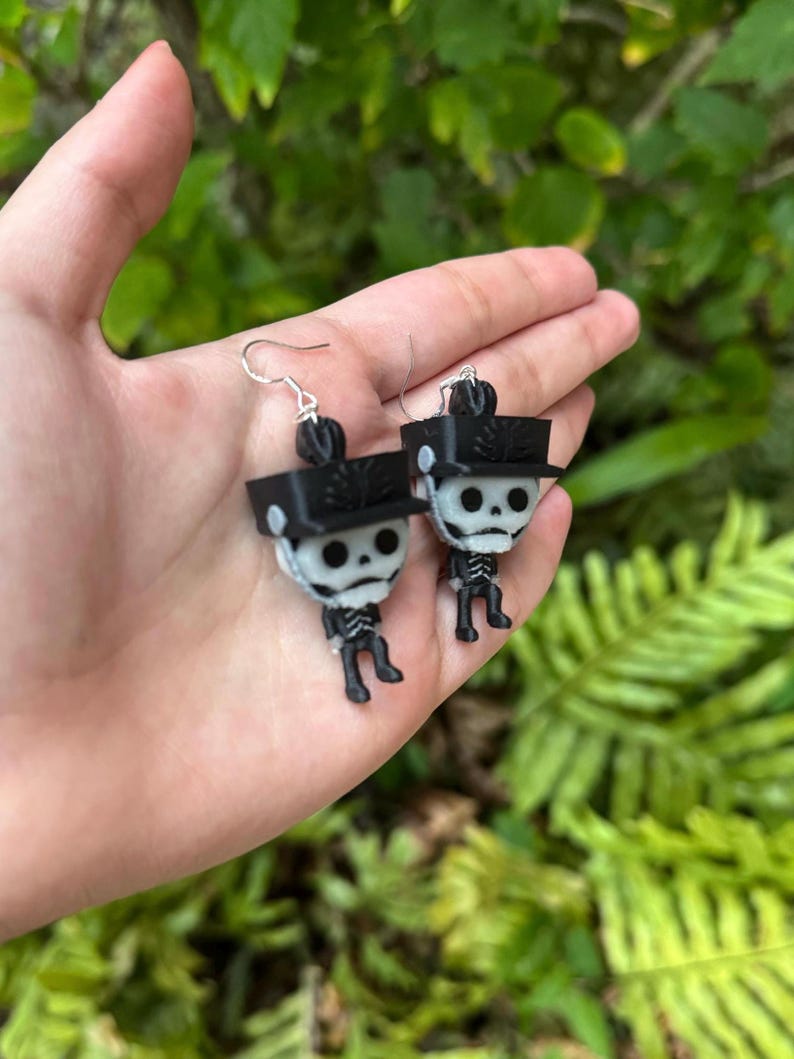 May include: A pair of black and white skeleton earrings with black hats. The earrings are hanging from silver hooks.