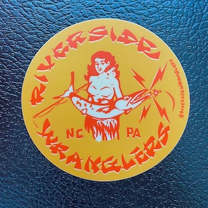Puede incluir: Una pegatina redonda con fondo amarillo y texto y gráficos naranjas. El texto dice "Riverside Wranglers" en una fuente curva. El gráfico es de una mujer sosteniendo un pez. La pegatina también incluye "NC" y "PA".