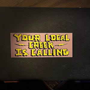 Puede incluir: Una pegatina rosa y amarilla con el texto "Your Local Creek Is Calling" en una fuente de dibujos animados.
