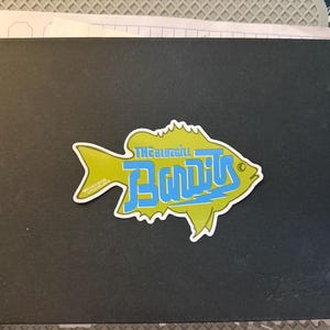 Puede incluir: Una pegatina de pez amarilla con el texto "The Bluegill Bandits" en letras azules. La pegatina está en una libreta negra.