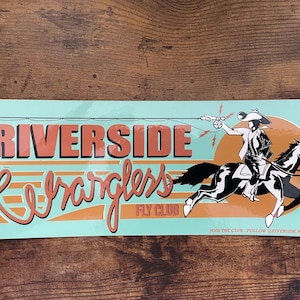 Puede incluir: Adhesivo rectangular con las palabras "Riverside Wranglers Fly Club" en naranja y blanco. El adhesivo presenta un vaquero a caballo pescando con mosca, con un fondo azul claro y una superficie de madera marrón.