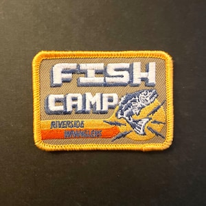 May include: A beige embroidered patch with yellow stitching. The patch features the text "FISH CAMP" in white with blue outlines. Below the text is a blue and white fish illustration with orange and yellow accents. The text "RIVERSIDE WRANGLERS" is in orange and white on a yellow stripe.