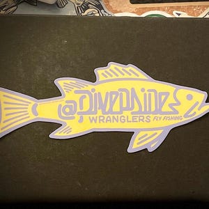 Puede incluir: Pegatina amarilla en forma de pez con el texto "La Divpsine" y "WRANGLERS FLY FISHING" en letras moradas.