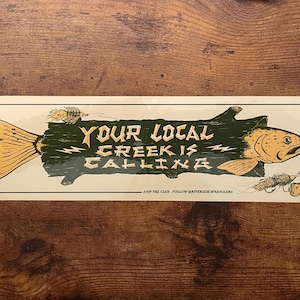 Puede incluir: Adhesivo rectangular con una ilustración de un pez. El pez es verde y dorado, con el texto "YOUR LOCAL CREEK IS CALLING" en una fuente estilizada. El adhesivo tiene un borde blanco y está sobre una superficie de madera.