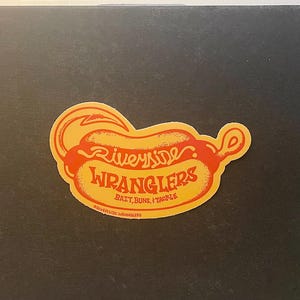 Puede incluir: Pegatina amarilla y roja con el texto "Riverside Wranglers" y las palabras "Bait, Buns, & Tackle" debajo. La pegatina tiene forma de hot dog con un gancho en el extremo.