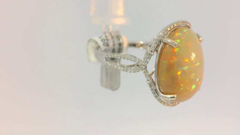 May include: A white gold ring with a large oval opal gemstone surrounded by a halo of small diamonds. The ring has a delicate, intricate design.