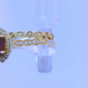 May include: A gold ring with a large oval-shaped red gemstone surrounded by smaller diamonds. The ring is set on a white background.