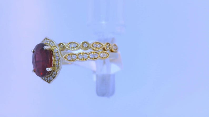 May include: A gold ring with a large oval-shaped red gemstone surrounded by smaller diamonds. The ring is set on a white background.