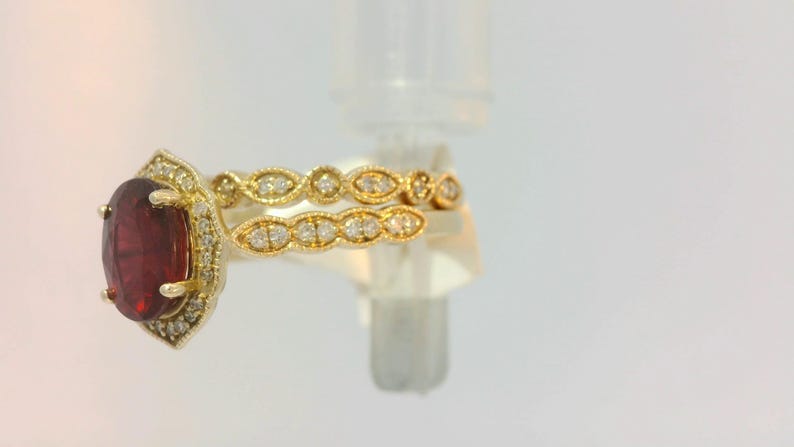May include: A gold ring set with a large oval-shaped red gemstone surrounded by smaller diamonds. The ring is paired with a matching band featuring a row of diamonds set in a delicate design.