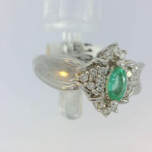 Pu&ograve; includere: Un anello d'argento con uno smeraldo verde circondato da diamanti pi&ugrave; piccoli. L'anello ha un design floreale delicato.