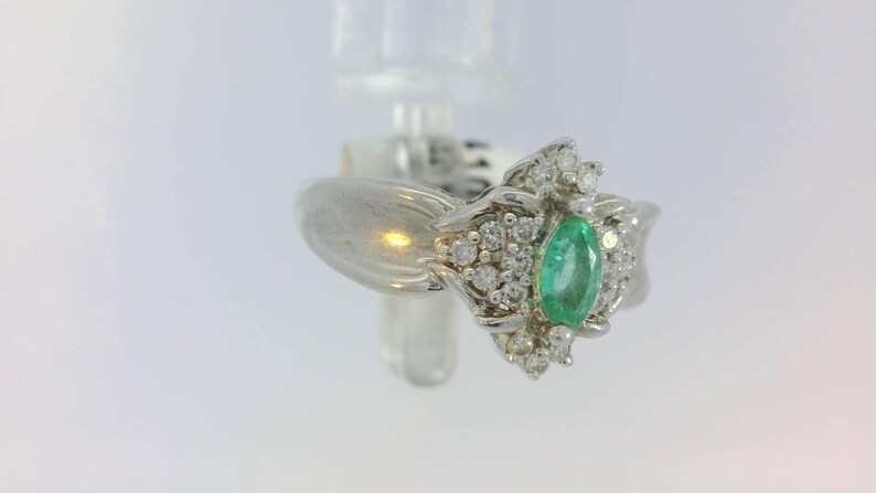 Pu&ograve; includere: Un anello d'argento con uno smeraldo verde circondato da diamanti pi&ugrave; piccoli. L'anello ha un design floreale delicato.