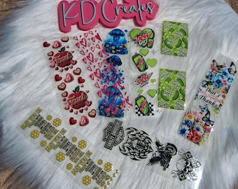 May include: A collection of clear stickers with various designs, including hearts, apples with the word "Teach", jellyfish, soda cans, pickles, butterflies, and the text "May you be blessed". A pink sign says "KD Creates".