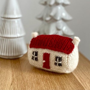 Könnte beinhalten: Ein gestricktes weißes Haus mit rotem Dach und schwarzen Fenstern. Das Haus steht auf einem Holztisch vor zwei weißen Keramik-Weihnachtsbäumen.