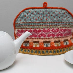 May include: A knitted tea cozy with a colorful geometric pattern in shades of blue, pink, orange, and brown. The cozy has a loop handle and is made with a variety of textures.
