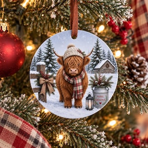 May include: A round ceramic Christmas ornament featuring a fluffy brown Highland cow wearing a beige knit hat and a red and green plaid scarf. The scene includes a snowy landscape with pine trees, a wooden fence, a lantern, and a metal bucket.