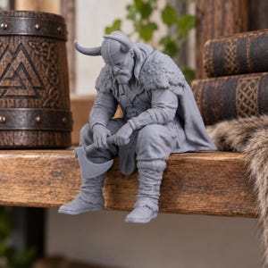 Könnte beinhalten: Eine graue, monochrome Figur eines sitzenden Wikingerkriegers mit gehörntem Helm und strukturiertem Umhang. Die Figur ist nach vorne gebeugt, trägt einen Bart und hält eine Streitaxt in beiden Händen, wobei der Axtkopf auf der Fläche ruht.