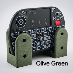 May include: An olive green keyboard holder with a black mini keyboard. The keyboard has white keys with blue lettering and a red button. The holder has two vertical supports with holes. The text "Rii x8" is visible on the keyboard.