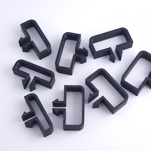 Può includere: Otto clip di plastica nera, ciascuna con un telaio rettangolare e una sporgenza a forma di T. Disposte su uno sfondo bianco, evidenziano il loro design. L'elemento a forma di T suggerisce una funzione o un meccanismo di fissaggio specifico. Realizzate in plastica.