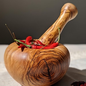 Könnte beinhalten: Ein hölzernes Mörser- und Stößel-Set gefüllt mit roten Chilischoten. Der Mörser ist rund mit breitem Boden, der Stößel lang und gebogen. Getrocknete rote Chilischoten liegen um den Mörser herum.