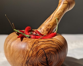 Könnte beinhalten: Ein hölzernes Mörser- und Stößel-Set gefüllt mit roten Chilischoten. Der Mörser ist rund mit breitem Boden, der Stößel lang und gebogen. Getrocknete rote Chilischoten liegen um den Mörser herum.