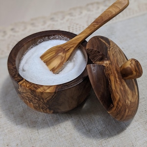 May include: A wooden salt cellar with a lid and a small wooden spoon. The cellar is filled with white salt.