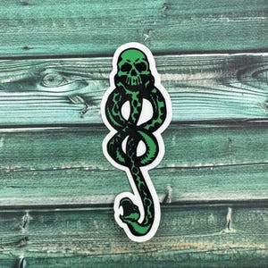 May include: Green and black sticker of the Dark Mark, a symbol from the Harry Potter series. The symbol is a snake with a skull at the top.