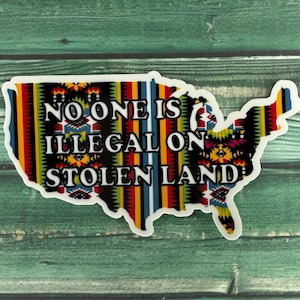 May include: A sticker with a colorful, geometric pattern in the shape of the United States. The sticker has the text "NO ONE IS ILLEGAL ON STOLEN LAND" in white letters.