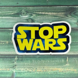 May include: A white sticker with a yellow "STOP WARS" text on a blue galaxy background. The sticker is on a green wooden surface.