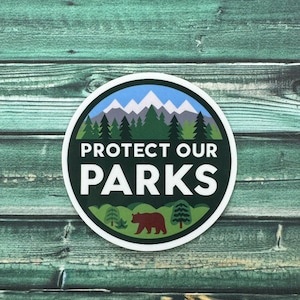 May include: A round sticker with a green background and white text that says "Protect Our Parks". The sticker features a mountain range with snow-capped peaks, a brown bear, and green trees.