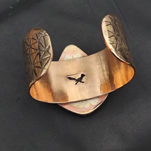 May include: A copper-coloured cuff bracelet with geometric patterned ends. The bracelet features a central, diamond-shaped design with a small, black roadrunner silhouette. The cuff is open-backed.