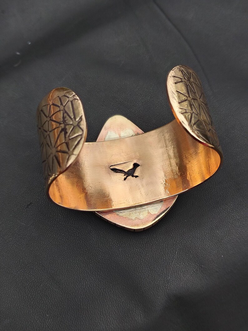 May include: A copper-coloured cuff bracelet with geometric patterned ends. The bracelet features a central, diamond-shaped design with a small, black roadrunner silhouette. The cuff is open-backed.
