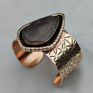 May include: A gold-coloured cuff bracelet featuring a large, teardrop-shaped stone. The stone is dark grey with a textured surface, set in a gold frame. The cuff has a geometric pattern.